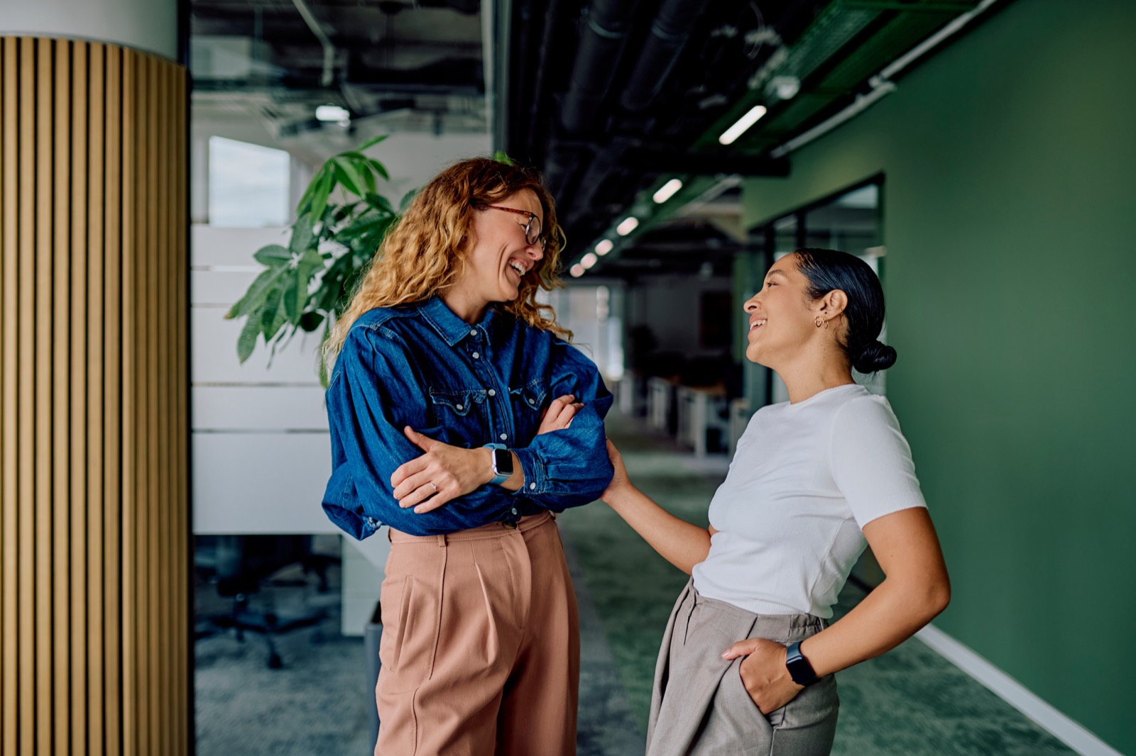 Two colleagues laughing together in a bright office hallway beside a green wall and plants