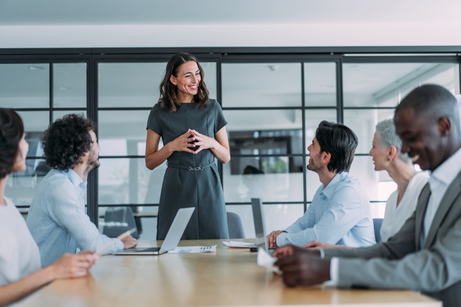 A team leader standing at the head of a meeting table, smiling as her colleagues talk with her