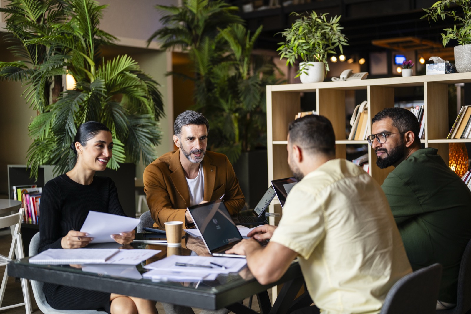A team of four working together at a table with laptops and papers in a bright office with plants