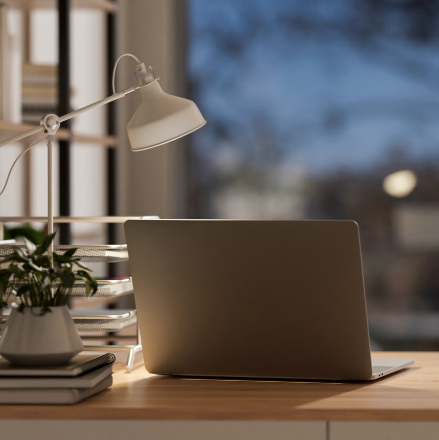 A laptop open on a desk at dusk, lit by a desk lamp.